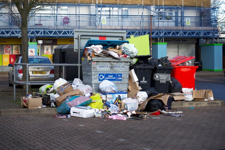 A collection of overflowing rubbish bins and scattered waste on a paved urban street corner. The scene includes multiple black and red trash bags, flattened cardboard boxes, and paper or plastic packaging strewn across the ground and atop grey and black communal recycling containers labeled for mixed paper and cardboard. In the background, a grey utility box and a silver car parked adjacent to a metal railing are visible, with a building featuring shopfronts and signage further behind. The area is illuminated by natural daylight, and the scene suggests a temporary accumulation of waste awaiting collection, reflecting issues related to private rubbish disposal and alternative waste handling outside of municipal services, with a focus on rubbish removal efforts.
