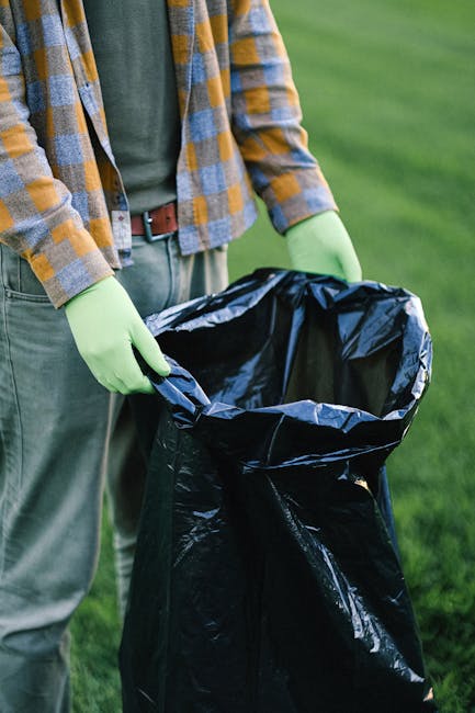 A person standing outdoors on a grassy area, wearing a plaid shirt over a green top and grey trousers, is holding open a large black rubbish bag lined with a metallic inner layer. The individual is wearing light green rubber gloves, indicating they are engaged in waste collection or disposal. The scene suggests an environmental clean-up activity, with the focus on the rubbish bag likely used for collecting waste materials. The background features a well-maintained lawn with vibrant green grass, and the lighting appears natural, possibly on a clear day. The image captures the process of private waste handling or an alternative rubbish collection at a community or park setting, which may relate to local rubbish removal services such as those offered by rubbishremovalstjohnswood.com, emphasizing on-site clearance or independent collection practices.