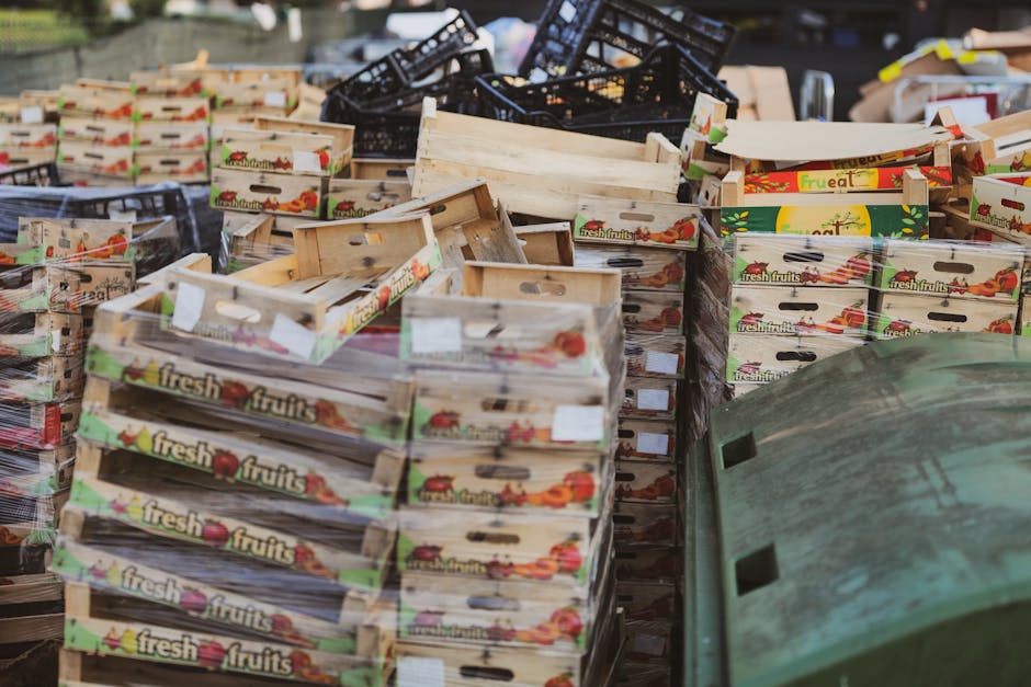 A large outdoor scene featuring numerous wooden crates labeled 'fresh fruits' with colourful fruit illustrations and transparent plastic wrapping, stacked haphazardly in a cluttered area. The crates are arranged in various directions, some leaning on each other, with a mix of empty and partially filled boxes showing wooden textures and natural light brown finishes. Behind the crates, there are black plastic fruit crates and other miscellaneous packaging materials, creating a chaotic backdrop. To the right, a large, green, slightly weathered plastic bin with a textured surface and a rectangular slot on its side is visible. The setting appears to be an outdoor storage or waste collection site on a paved surface, possibly a back area adjacent to a commercial or wholesale fruit supplier, with ambient daylight illuminating the scene. The overall environment suggests the handling or disposal of food packaging waste, aligning with independent waste collection or private disposal activities that a company like [COMPANY_NAME] might support as part of their rubbish removal services.