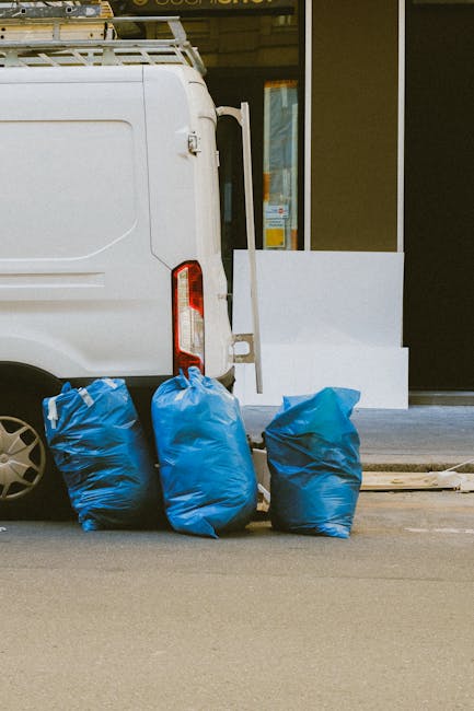 A person standing outdoors on a grassy area, wearing a plaid shirt over a green top and grey trousers, is holding open a large black rubbish bag lined with a metallic inner layer. The individual is wearing light green rubber gloves, indicating they are engaged in waste collection or disposal. The scene suggests an environmental clean-up activity, with the focus on the rubbish bag likely used for collecting waste materials. The background features a well-maintained lawn with vibrant green grass, and the lighting appears natural, possibly on a clear day. The image captures the process of private waste handling or an alternative rubbish collection at a community or park setting, which may relate to local rubbish removal services such as those offered by rubbishremovalstjohnswood.com, emphasizing on-site clearance or independent collection practices.