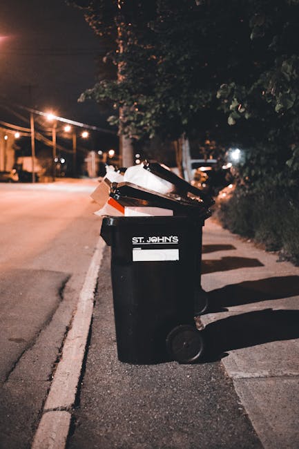 A black wheeled rubbish bin with a white and red reflective sticker on the side, positioned on a paved sidewalk next to a curb in a residential area during nighttime. The bin has an open lid revealing assorted waste materials including cardboard, paper, and plastic items. The background features a dimly lit street with warm streetlights, trees, and parked cars, suggesting an urban environment suitable for private waste collection or on-site rubbish removal services. The scene is captured under low-angle lighting, creating shadows on the pavement, and the bin bears the name 'St. John's,' indicating a local or community collection service. Surrounding elements include a tall hedge to the right and overhead power lines, with a dark sky overhead, emphasizing the quiet, evening atmosphere typical of a neighborhood receiving rubbish collection or clearance from independent waste management providers like rubbishremovalstjohnswood.com.