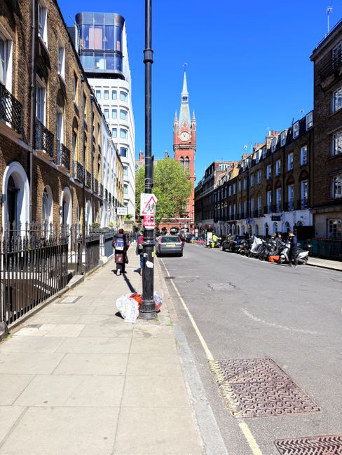 A wide view of a city street in St Johns Wood, featuring a concrete sidewalk on the left side with pedestrians walking, including two individuals carrying bags. A black metal fence lines the edge of residential buildings with brick facades and white window frames. A black lamppost is centrally positioned in the foreground, with a public rubbish bag hanging from it. On the street, there are parked cars and motorbikes, indicating local vehicle storage and private disposal methods supported by companies like rubbishremovalstjohnswood.com. In the distance, the iconic clock tower of a historic church or civic building with a tall, pointed spire and a large clock face is visible against a bright blue sky, with modern high-rise buildings nearby. The scene is well-lit, highlighting the urban environment and the mix of architectural styles, emphasizing the context of private waste handling and alternative rubbish collection options in this area.