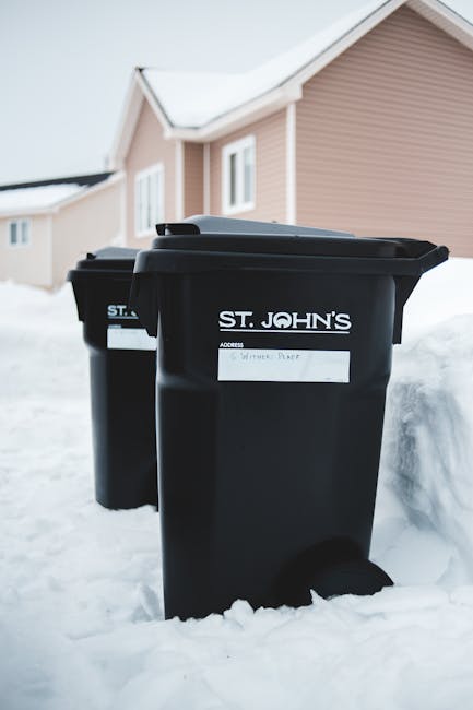 Two black rubbish bins with white labels on the front, labeled 'ST. JOHN'S' and designated for waste collection, are positioned on a snow-covered driveway in front of a residential building with light pink siding and white-framed windows. The bins are made of durable plastic with smooth textures and appear to have hinged lids. The scene is outdoors during winter, with snow surrounding the bins and partially covering the ground, indicating an environment where alternative waste management options, such as private collection or on-site clearance, may be utilized by residents rather than relying solely on local authority services. The background features a pitched roof and additional neighboring homes, suggesting a suburban setting. The scene’s natural lighting emphasizes the clarity of the bins' labels and the snow’s textured surface, contributing to a professional and neutral depiction suitable for waste management service content, as featured on rubbishremovalstjohnswood.com.